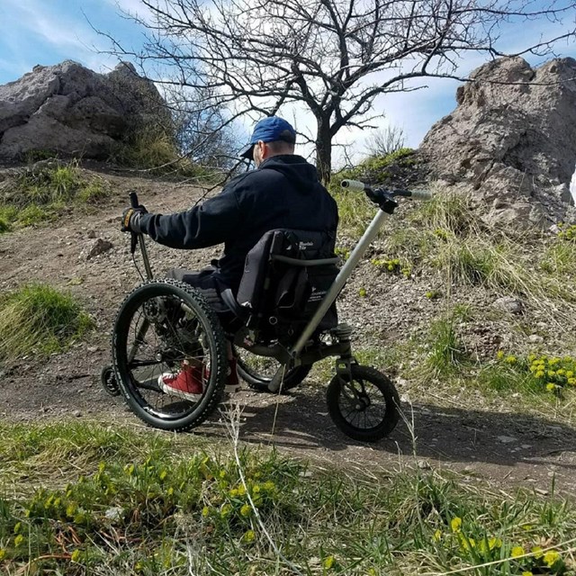 man pushing a levered manual wheelchair with sturdier tread, on a trail.  
