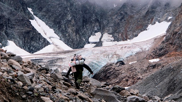 a person walks through a rocky scree field in front of a small valley glacier