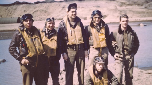 five pilots in military uniforms pose at the edge of a body of water.