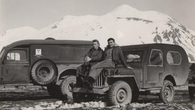 in a historic scene, two men sit on the hood of a jeep in front of a snow-covered mountain.