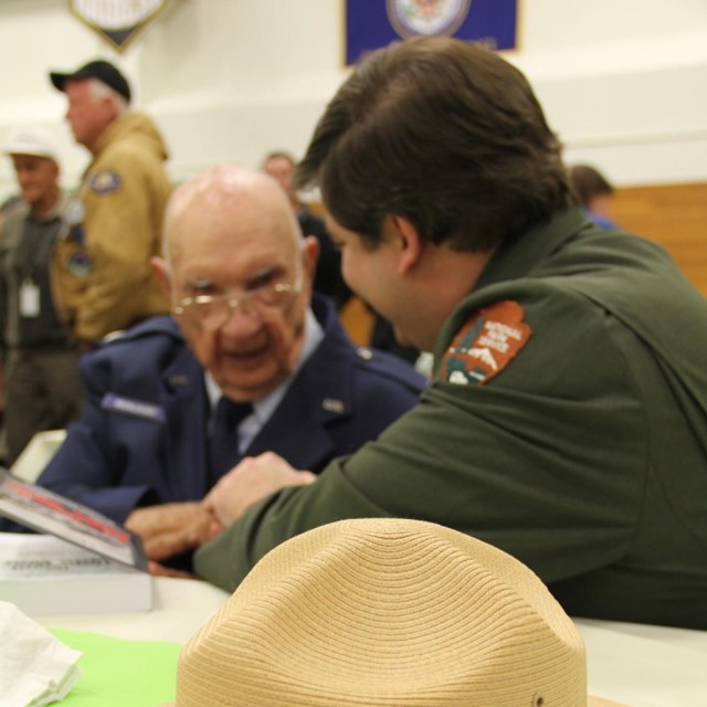 A man in a park ranger uniform speaks with an old man in a military uniform.