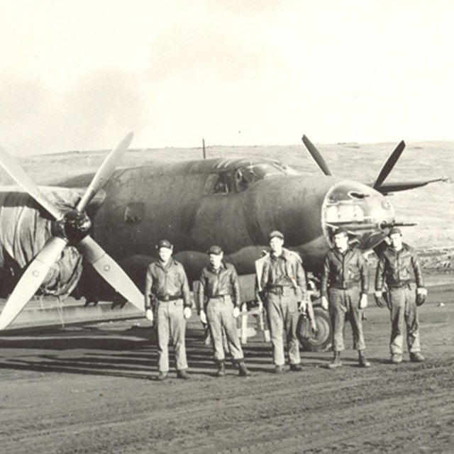 five uniformed servicemen walk on an airstrip in front of an airplane with two large propellers.