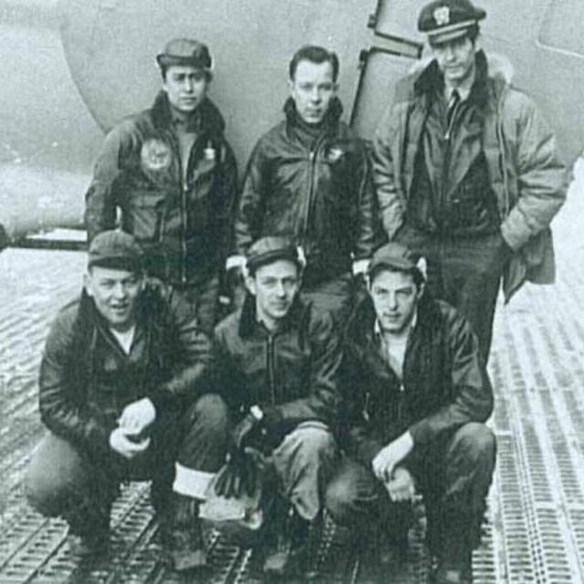 six men wearing leather uniforms pose in front of a plane.