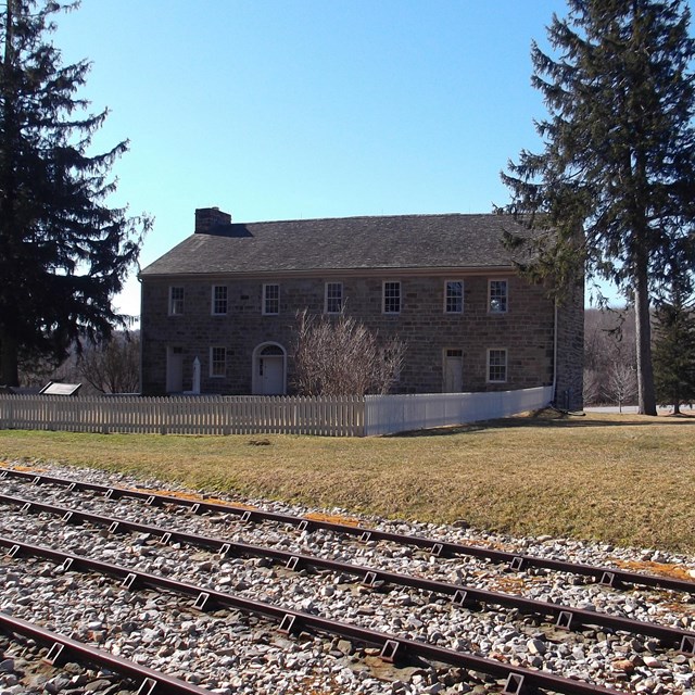 A large house in a grassy field behind train tracks, flanked by trees