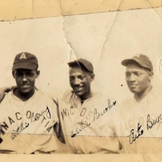 Three Black men in baseball uniforms stand arm in arm. 