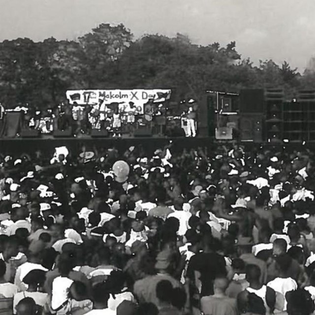 Thousands of people are shown in a crowd with a sign for Malcolm X Day.