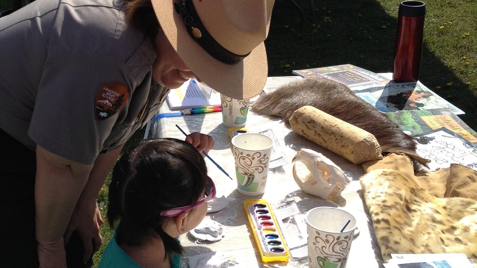 Outdoors; Park Ranger bending at hips to listen to a kid working at a table full of craft items.