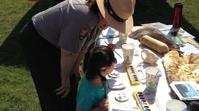Outdoors; Park Ranger bending at hips to listen to a kid working at a table full of craft items.
