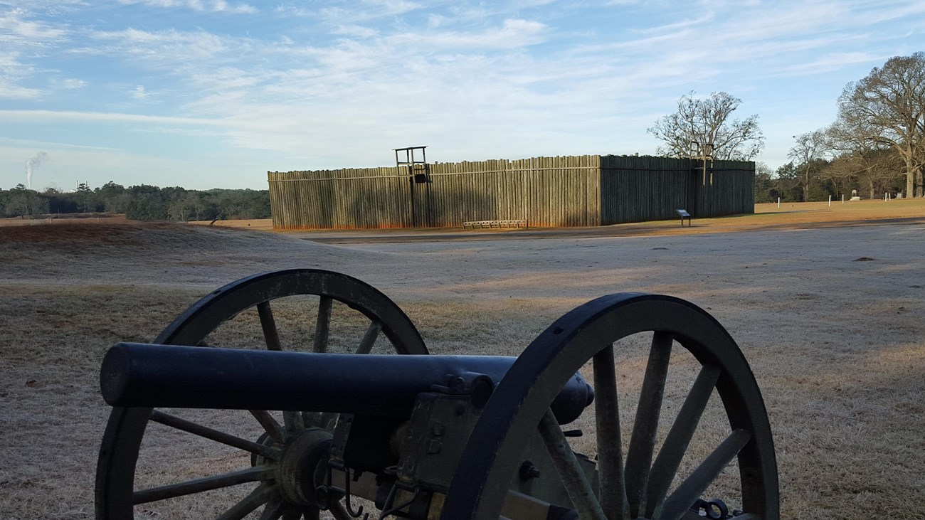 A cannon sits out in front of a replica of a wooden stockade wall