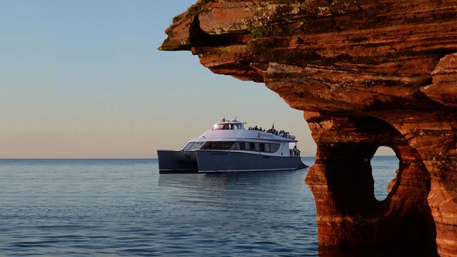 A large catamaran tour boat travels around an overhanging sandstone cliff at sunset.