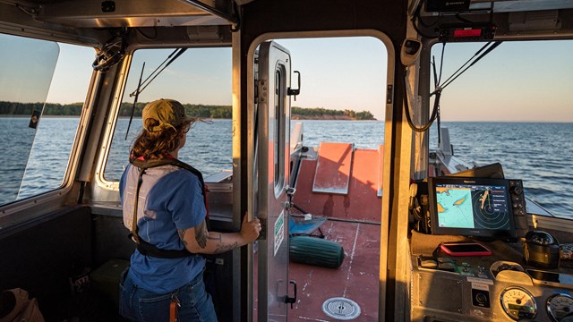 Photograph of a person standing inside a boat cabin looking out at the water during sunset.