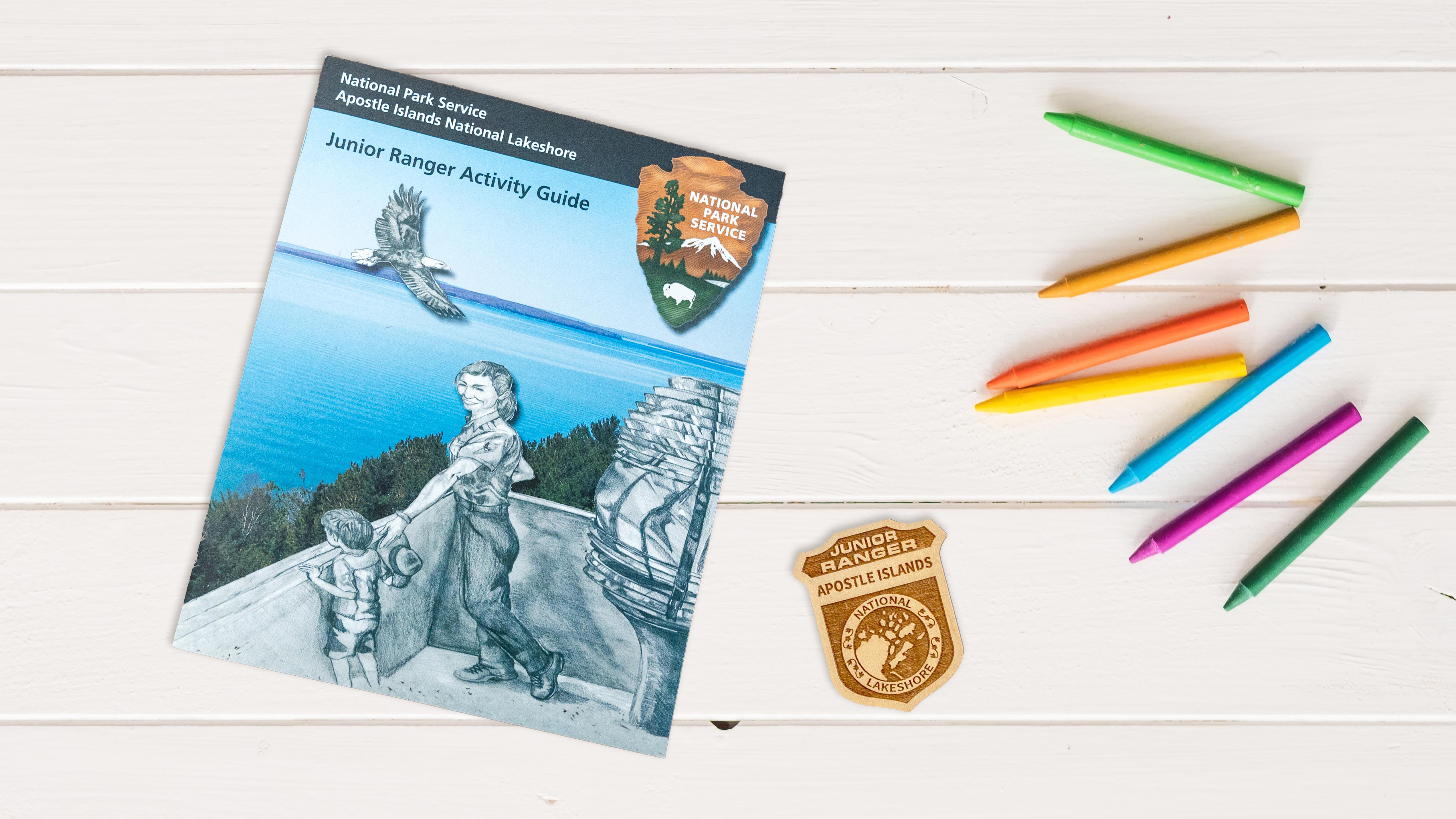 Photograph of Junior Ranger booklet, badge, and colorful crayons on white table.