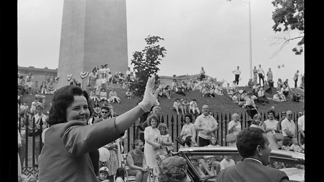 Black and white image of a woman waving from a car in front of the Bunker Hill Monument