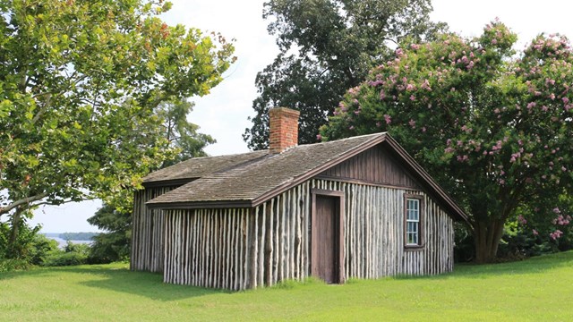 A brown wooden cabin with a chimney, surround by green trees and trimmed green grass.