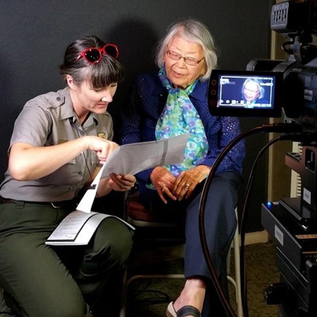 Two women look at a piece of paper in front of a camera