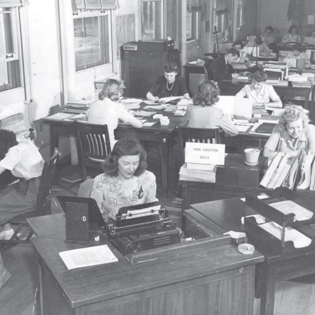 Black and white photo of women sitting at desks with lots of papers and type writers 