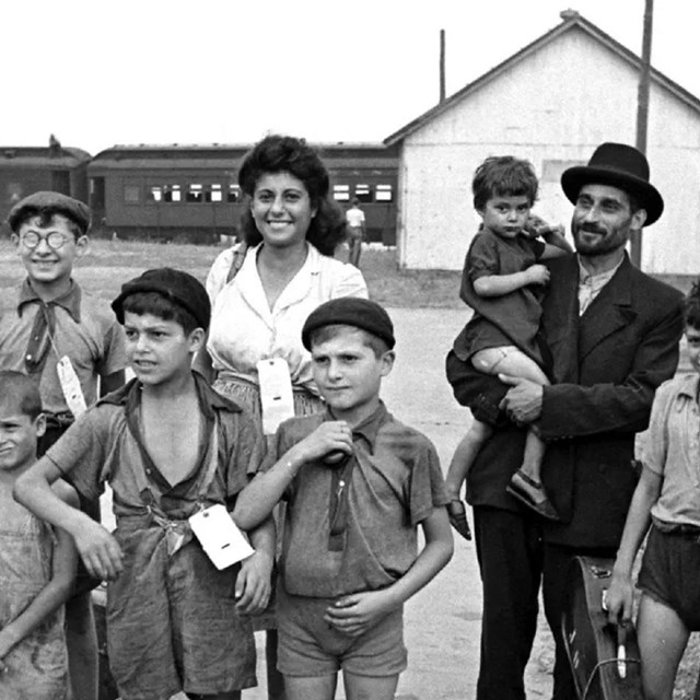 A group of men, women and children smile while standing in a yard in front of a low building.