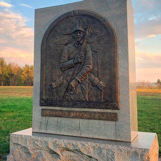 Stone monument with a metal engraving of a Black solider wearing a tricorn hat in a field