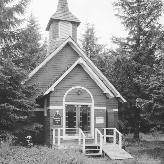Greyscale photo of small church between pine trees
