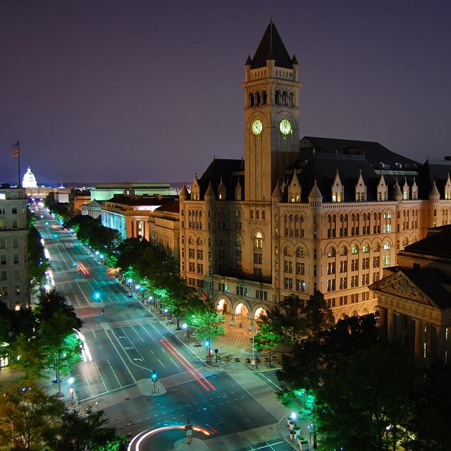 A large road a night. On one side is a large building with a bell tower.