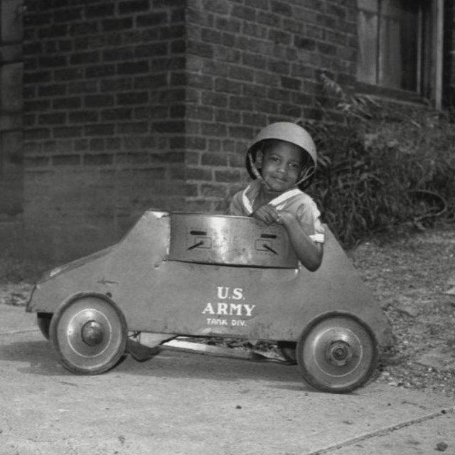 Black and white photo of an African American boy in a helmet in a toy car labeled US Army