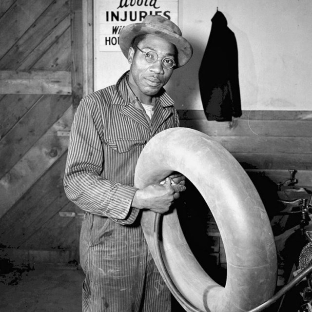 Black and white photo of an African American man in a hat and coveralls holding a tire
