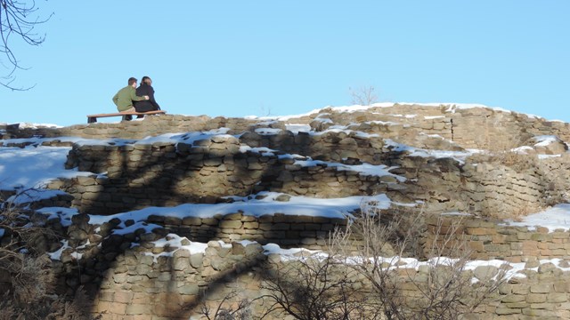 A view from behind of two visitors sitting on a park bench overlooking the ruins.