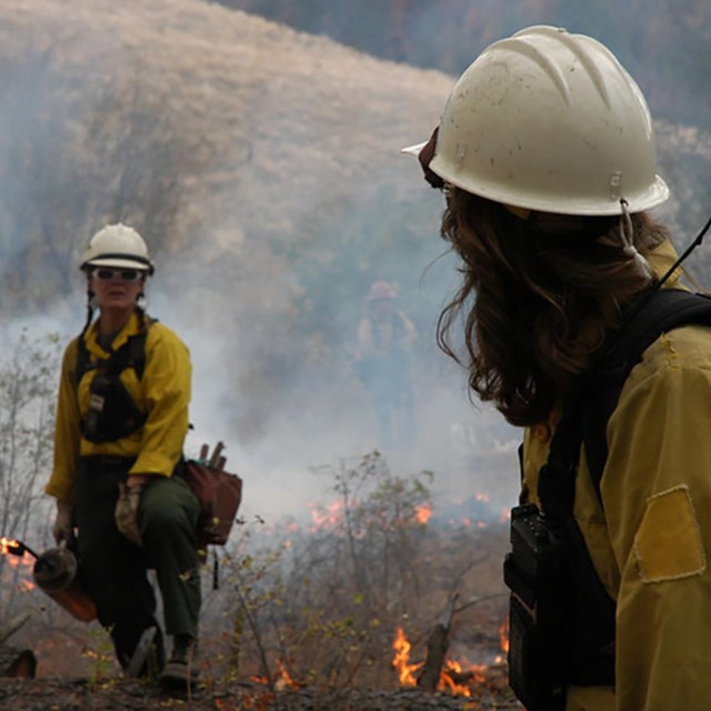 Two firefighters wearing yellow jackets and white helmets surrounded by smoke.