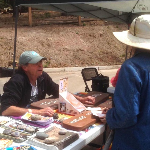 A man sits at a table filled with brochures and talks to a woman in a floppy hat.