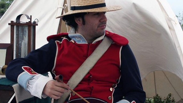 Male soldier in straw hat and red military uniform sits with pipe in hand