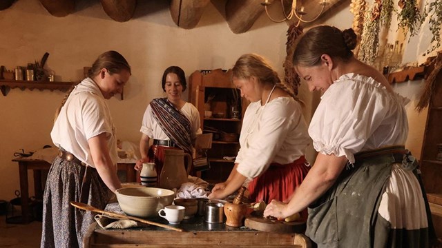 Women in historic costume work together in a kitchen with a stone hearth