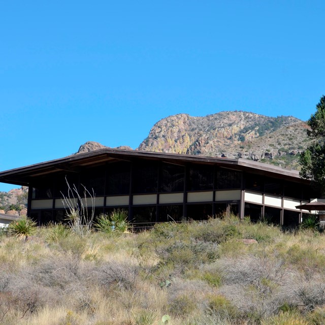 A low brown building sits on a hill with mountains in the background.