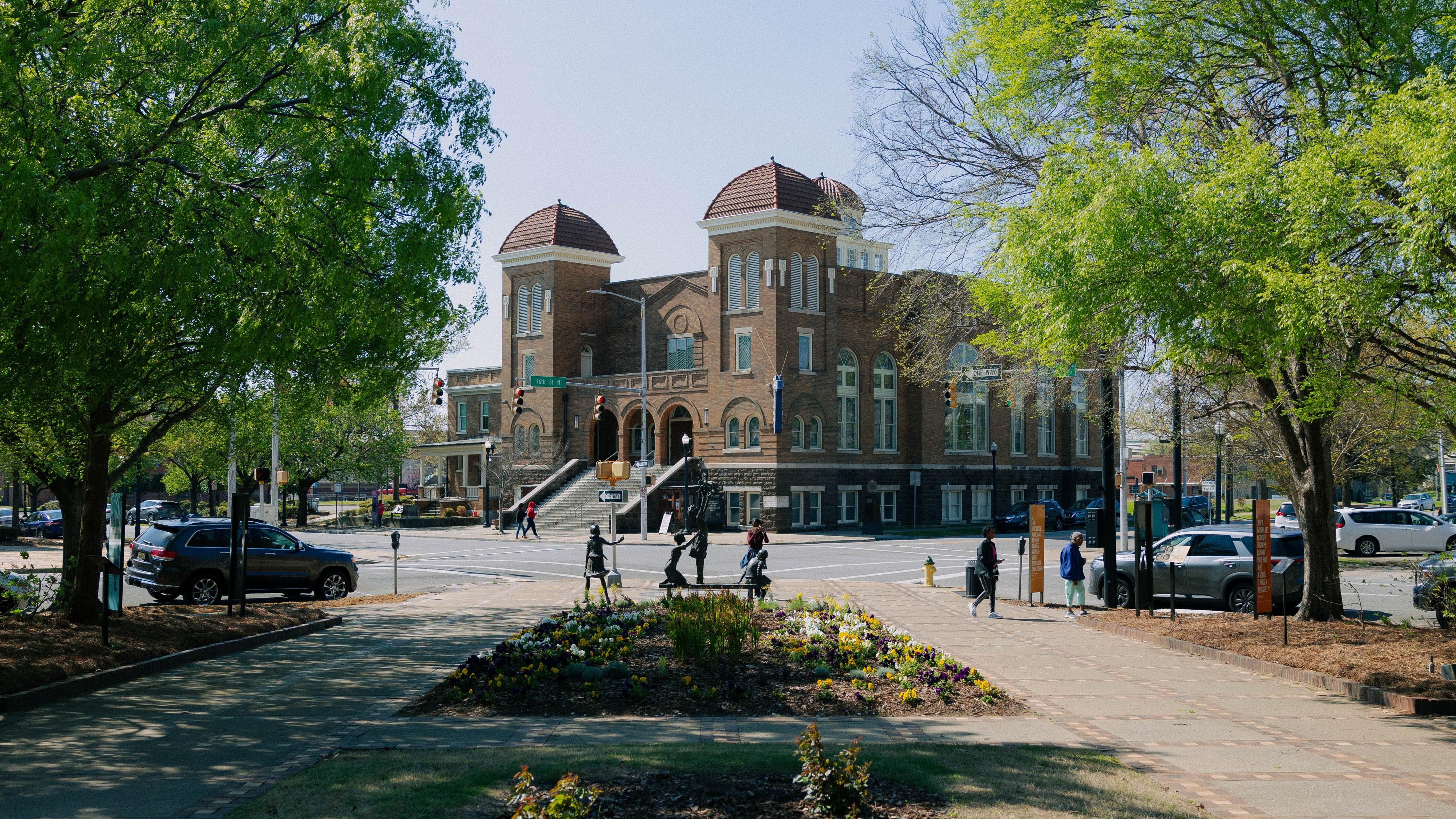 A large brick church with domes with a statue of 4 girls in a park.