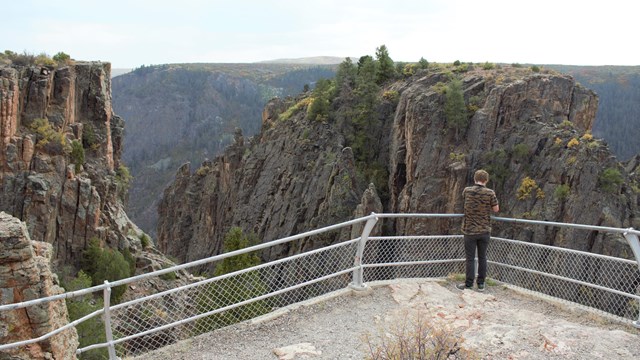 A person standing near a chain link fenced overlook by a canyon rim.