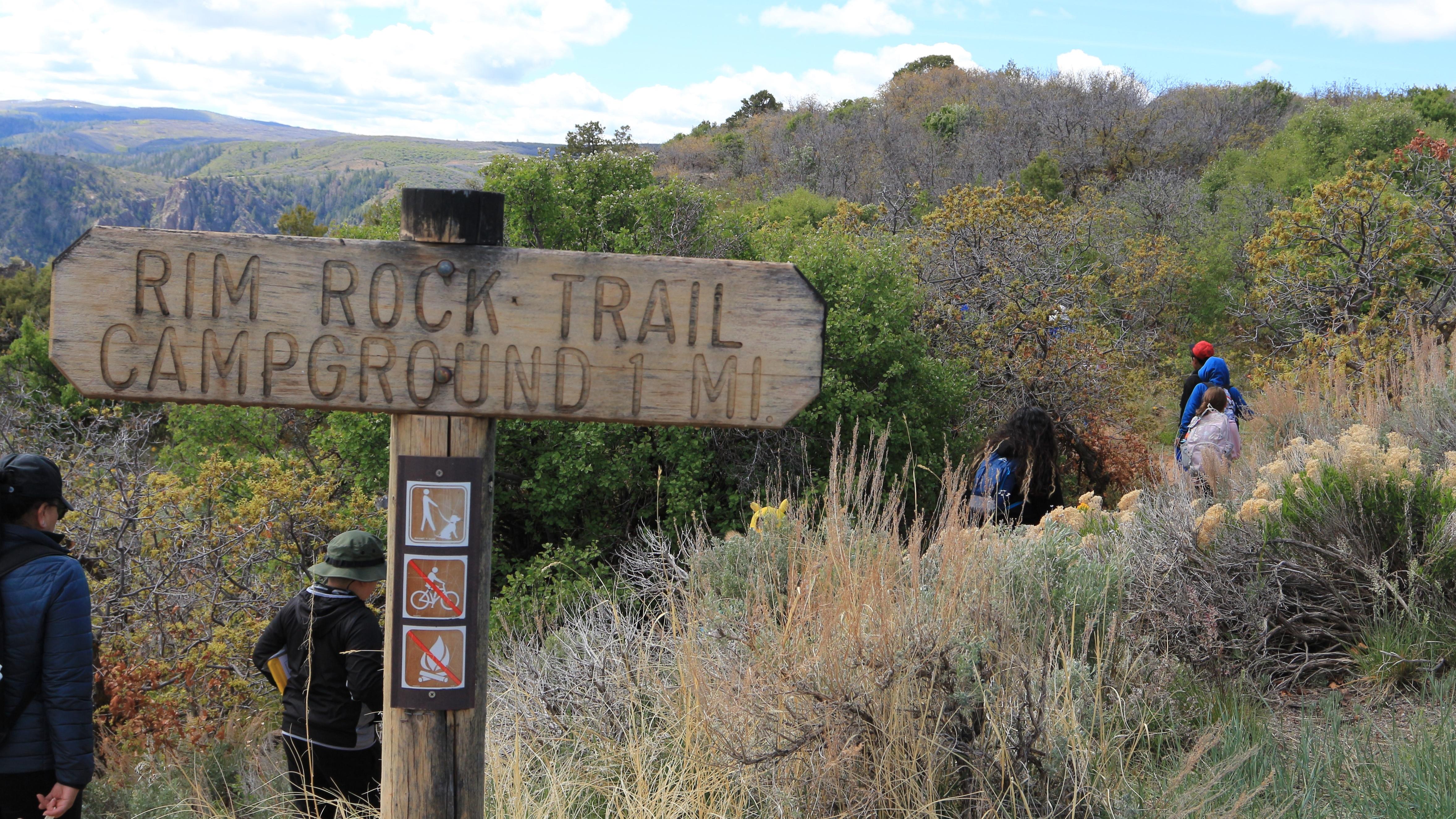 Adults and children with jackets and backpacks hike along a trail. Wooden trail sign in foreground.
