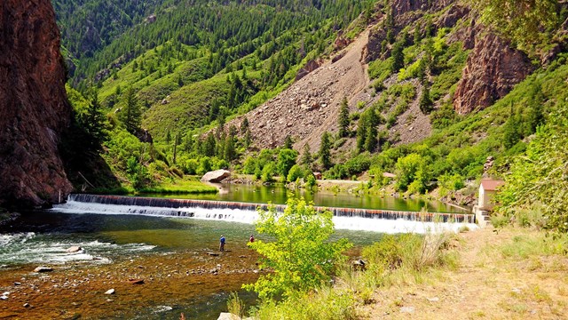 fisherman wading in the river in front of a short waterfall in the canyon