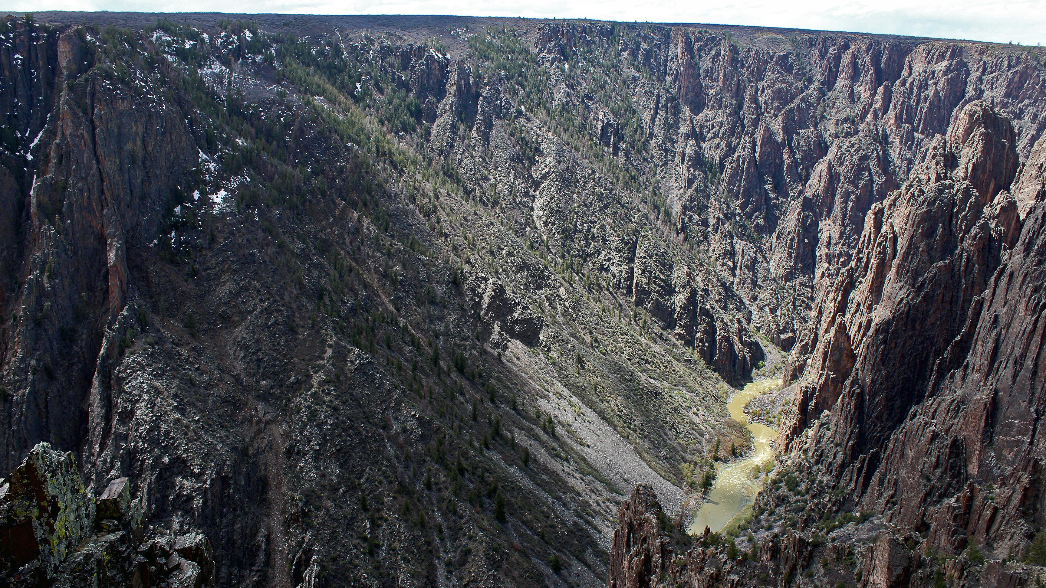 Broad view of a deep rocky canyon with cloud shadows and a greenish muddy river far below.