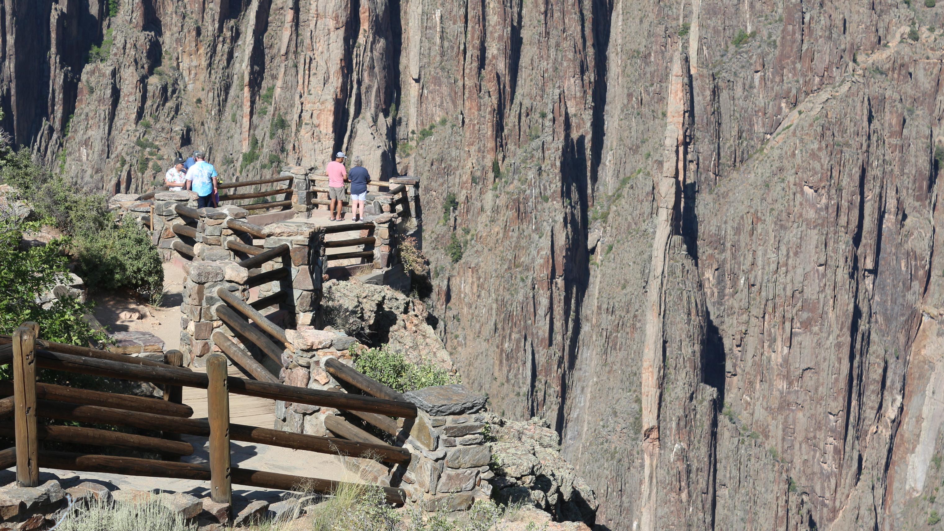 People standing at a fenced overlook at the edge of a canyon. Steep, canyon walls are beyond them.