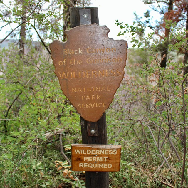A wooden arrowhead sign and smaller rectangular sign on a brown post