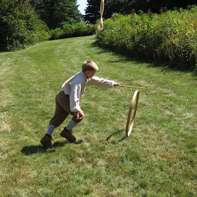 A child plays with a wooden stick and hoop in the grass.