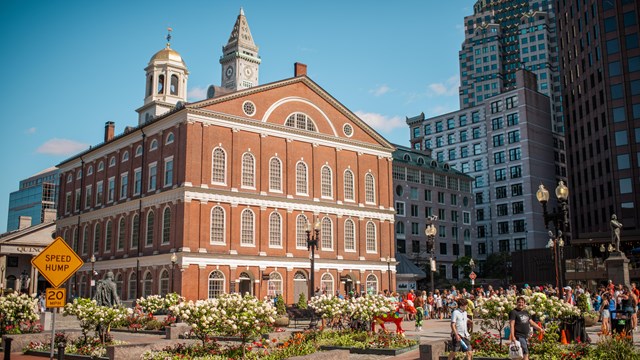 Faneuil Hall, a multi-story brick building with a cupola. In front are flower beds & people walking