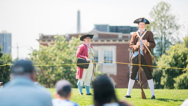 Two Park rangers dressed in period clothing, one has a musket to her side and speaks to the audience
