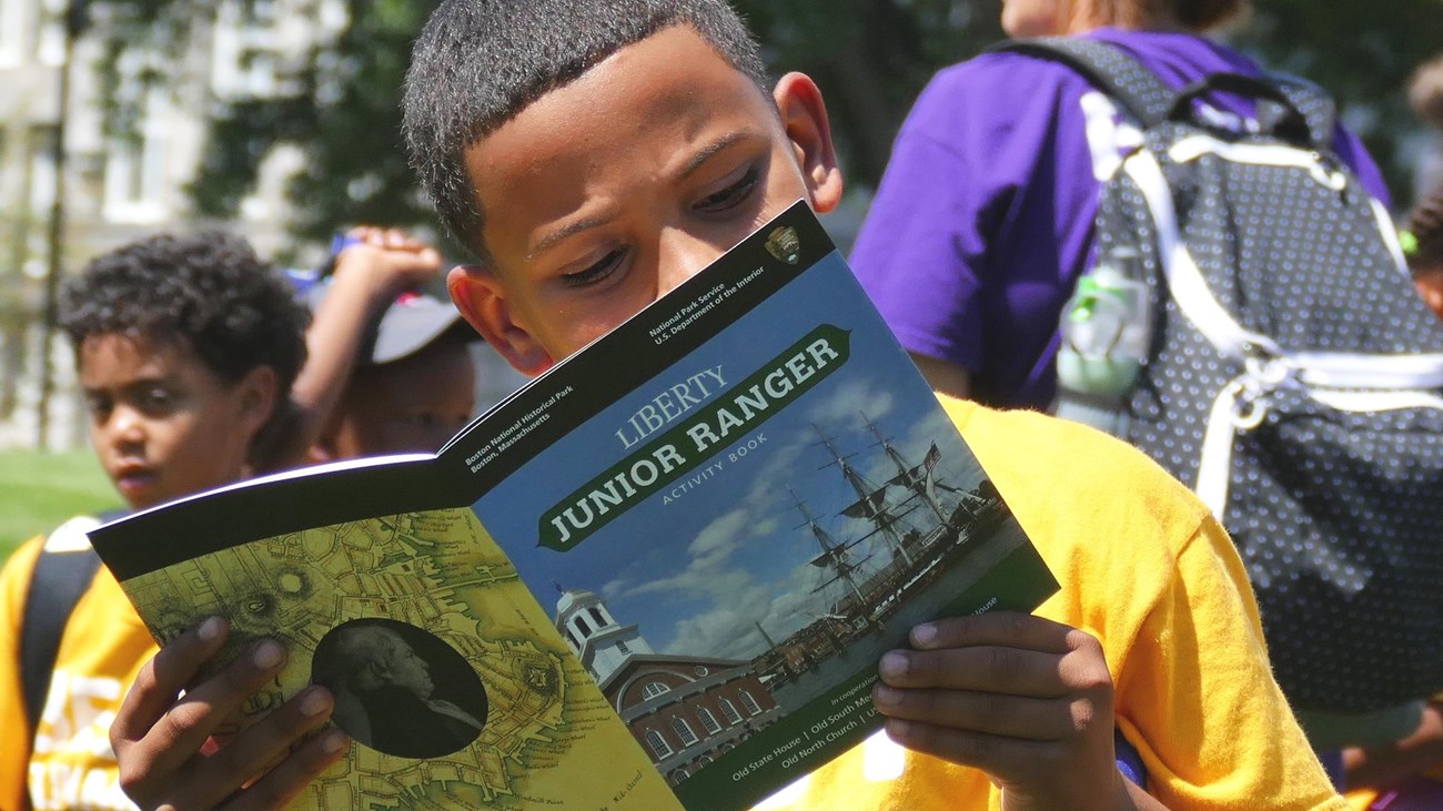 Child holding up a Junior Ranger book in front of his face and looking through it. 