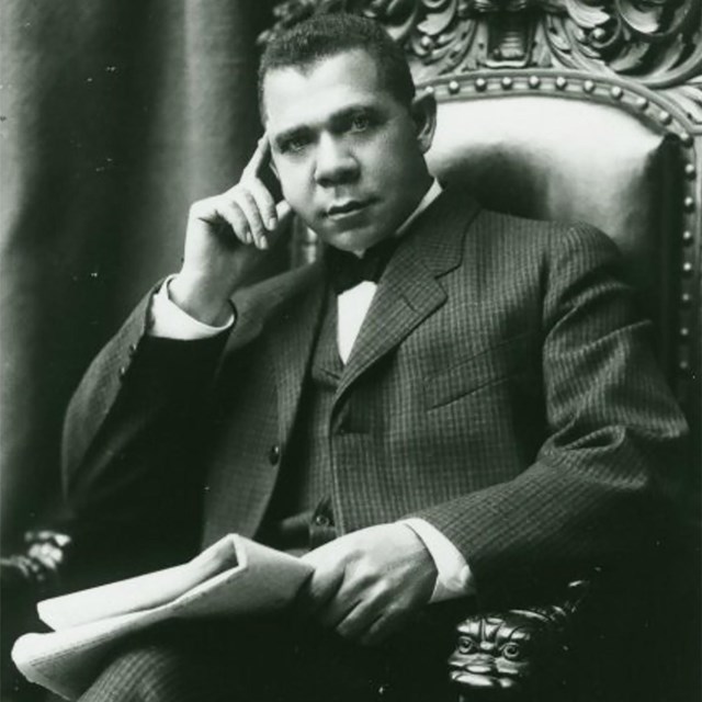 A photograph of Booker T. Washington sitting in a chair with a folded newspaper in his lap. 