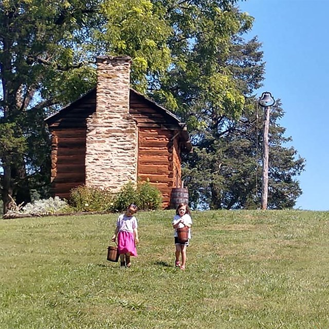 Two young children carrying wooden buckets down a hillside with a cabin in the background. 