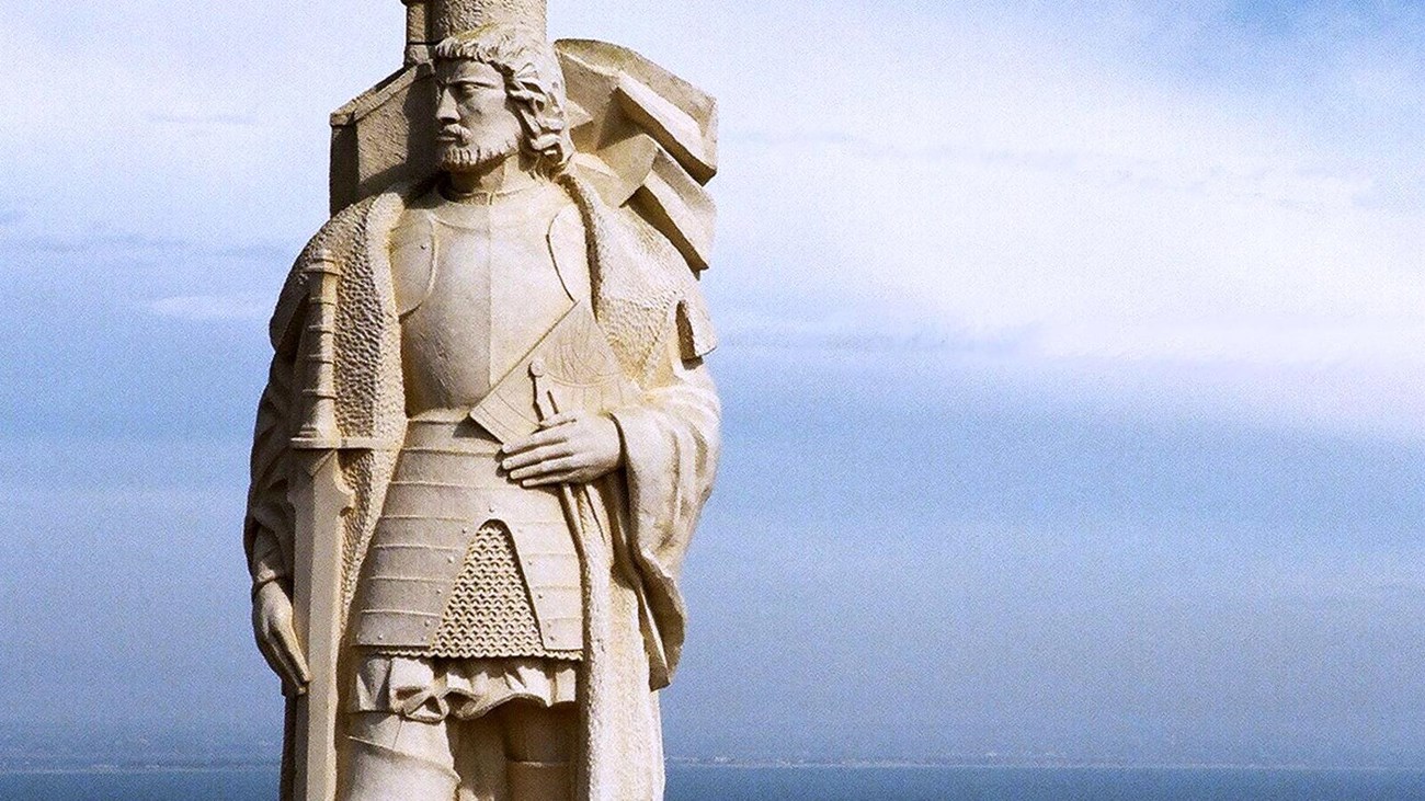 White stone statue of a man with cape. Blue ocean and blue sky with white clouds behind it.