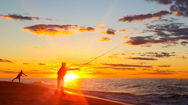 A glorious sun sets behind several fishermen casting into the crashing surf.