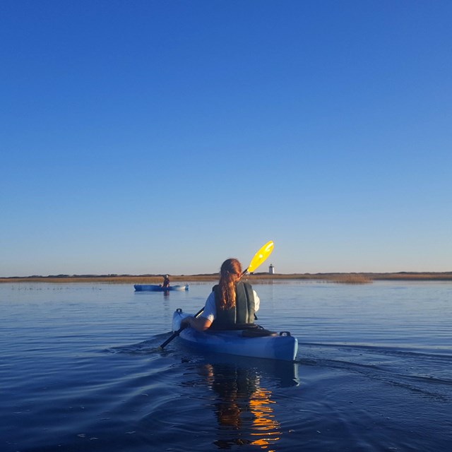 A ranger pulls a canoe down a ramp towards a pond, as a mother and son follow holding paddles.