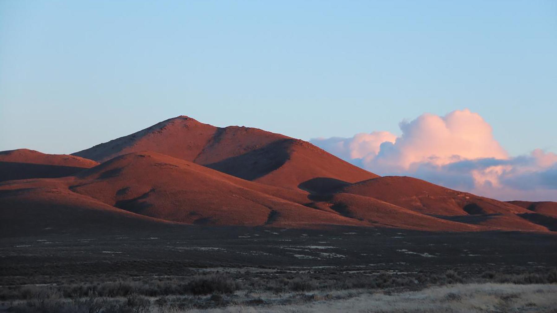 A large, smooth looking hill, colored red from the sunset, with distant billowing clouds.