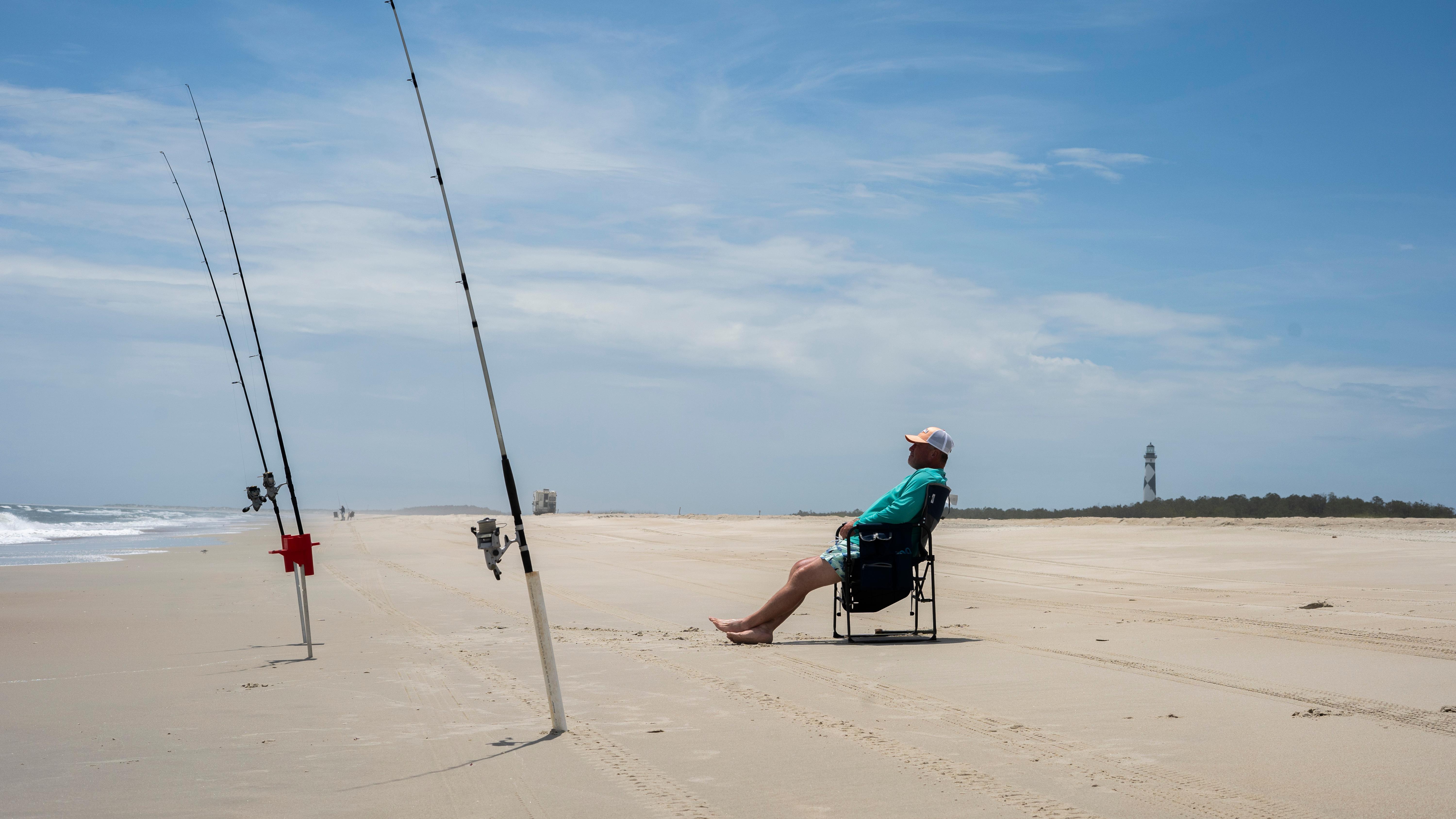 A angler sits on a chair with fishing poles in front of him. 
